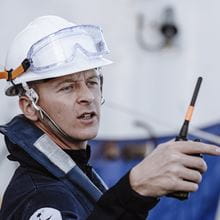 Sailor wearing a white hard hat and carrying a walkie talkie points 