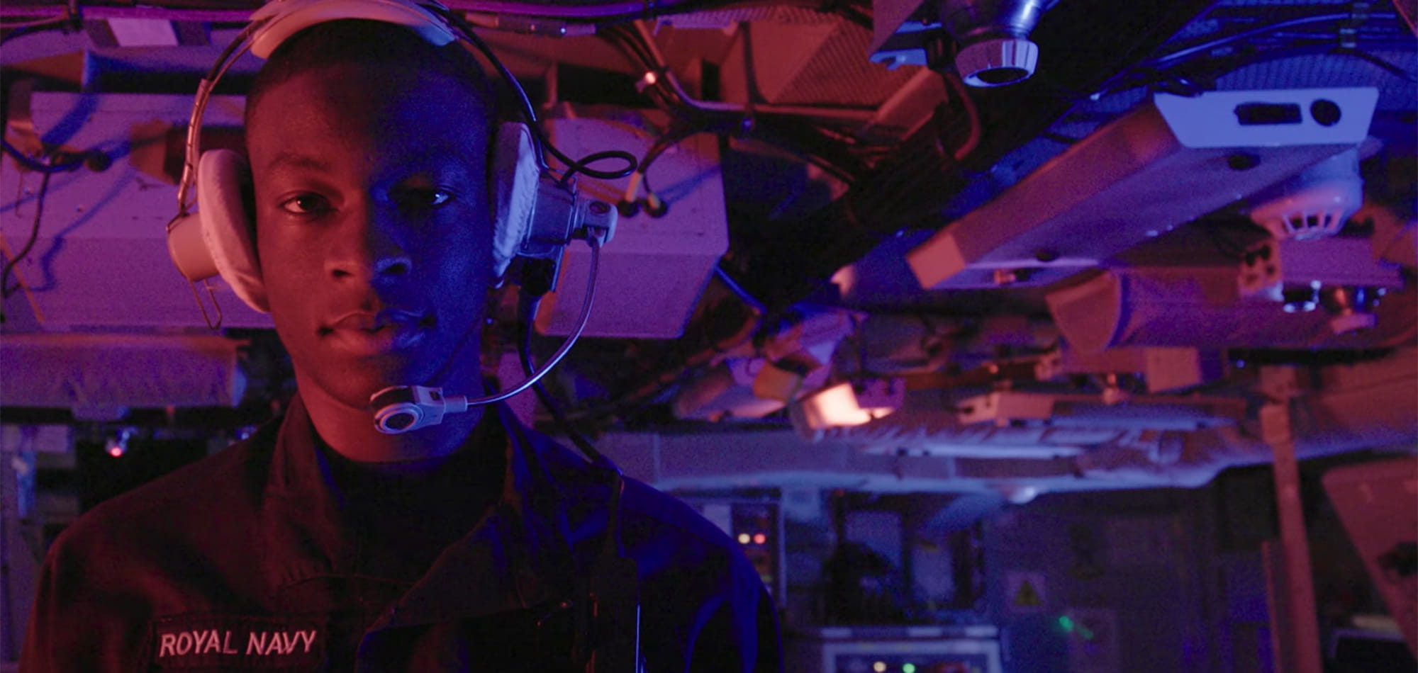 Royal Navy submariner wearing stands in a purple-lit control room