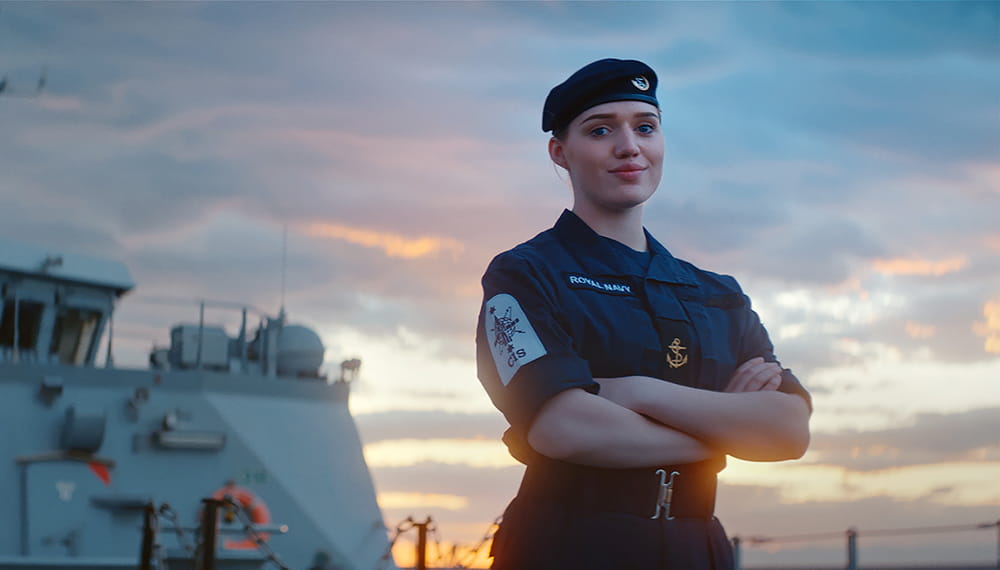 A uniformed crew member stands proudly on the deck of a ship during sunset