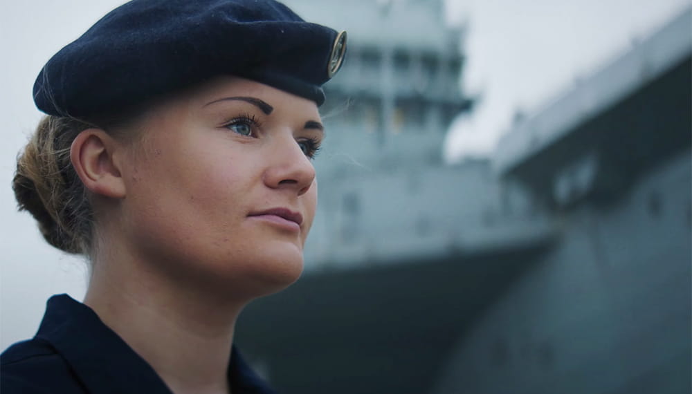 uniformed royal navy sailor with aircraft carrier in background looking into distance