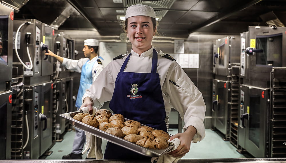 Royal Navy Chef looks at camera holding baking tray with potatoes.