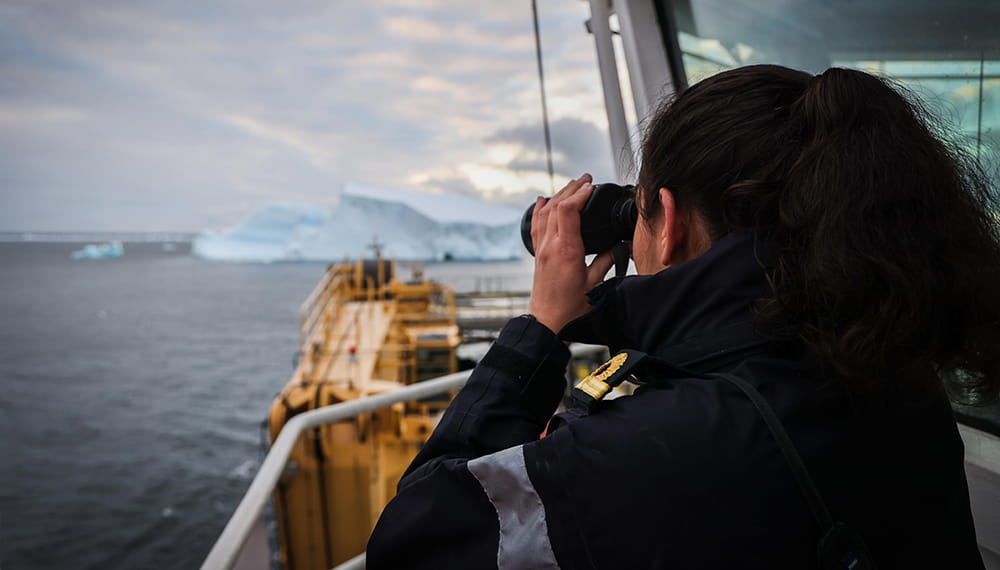 uniformed long brown haired officer outside bridge looking through binoculars with dark seas and iceberg in distance