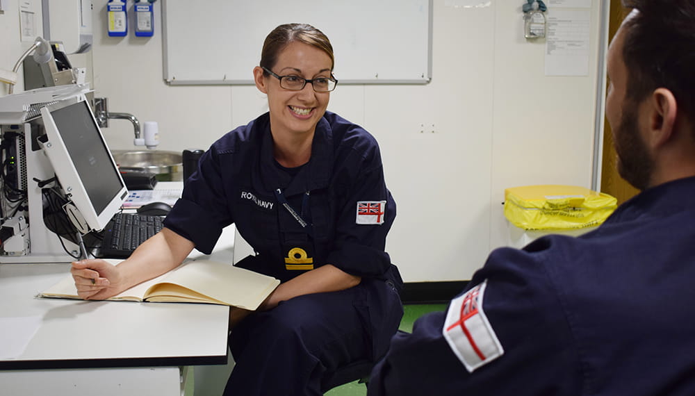 smiling dark blonde haired uniformed medical officer speaks with colleague in room with computer screen on desk to the left