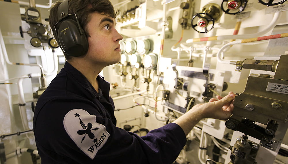 blue uniformed brown-haired weapons engineer submariner inspecting a panel in the bombshop surrounded by equipment