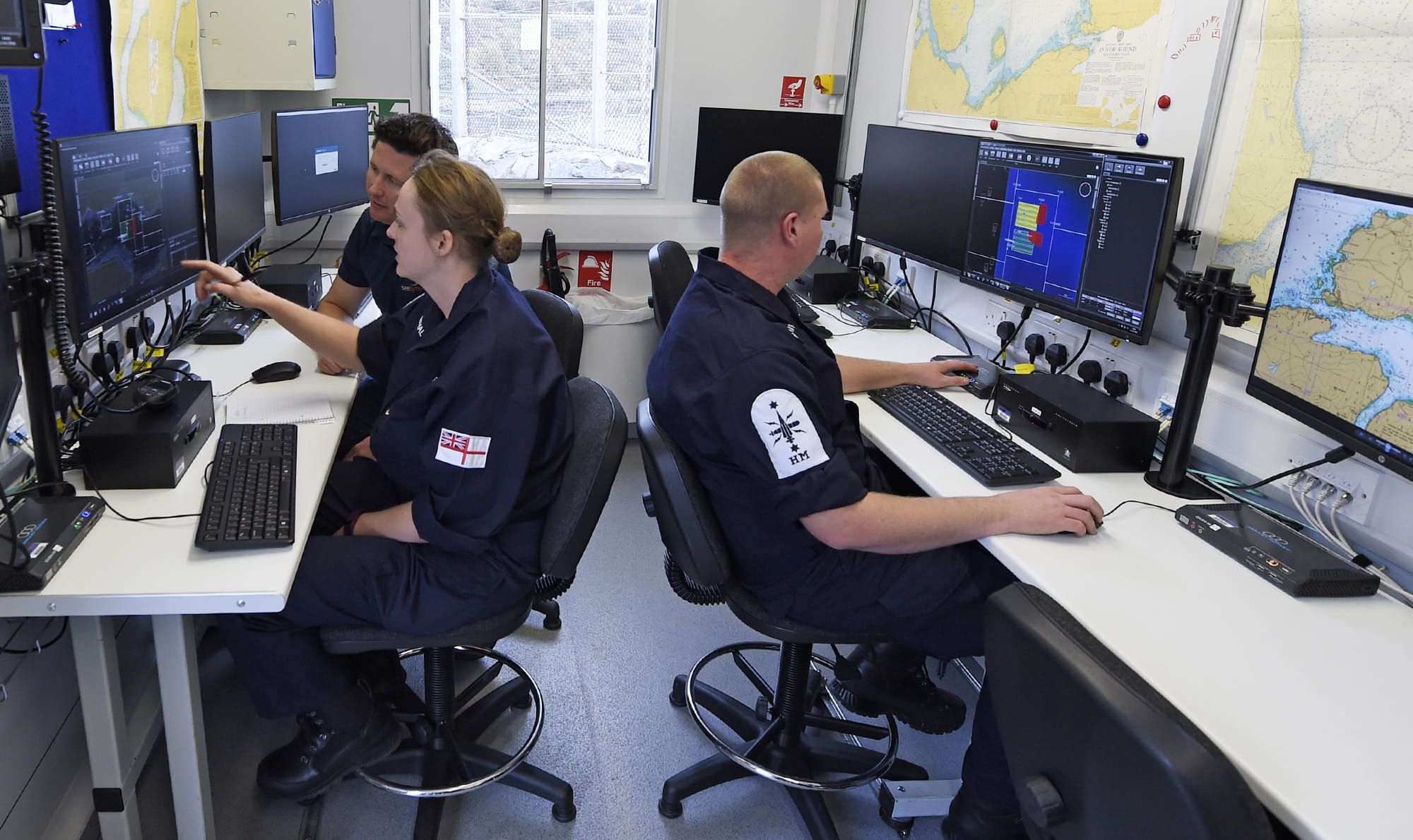 Group of Navy personnel working at desks with computers. The command operations module located at the British Underwater Test & Evaluation Centre (BUTEC) during Unmanned Warrior (UW) allows for the control and tasking of unmanned vehicles from multiple suppliers using a generic workstation. 