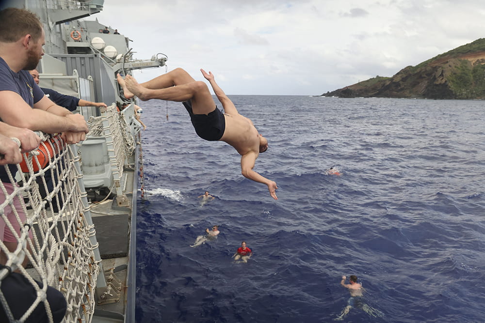 a member of the royal navy leaps off the side of a ship into the ocean with a light coloured sky and sloping forested hill in the background