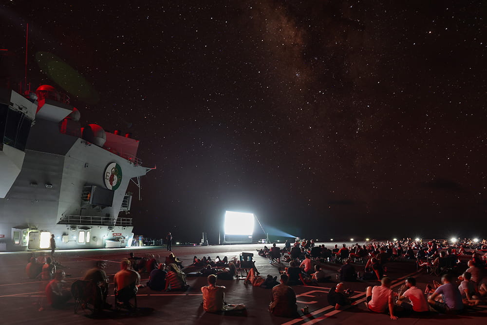 ship's company and embarked personnel enjoy a movie night on the flight deck under a starry sky