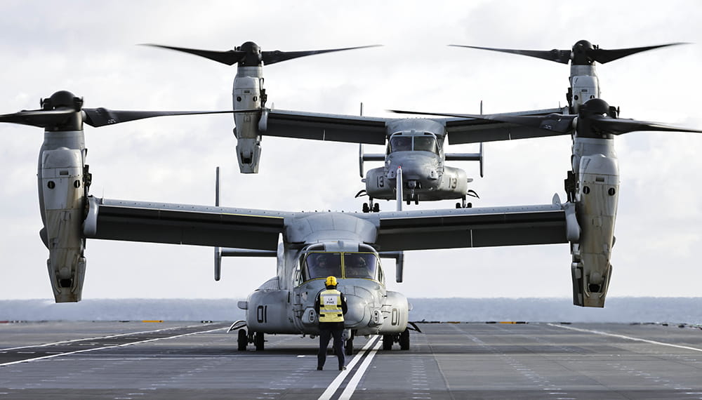 two grey V22 Osprey tiltrotor aircrafts land on deck of aircraft carrier HMS Prince of Wales with grey cloudy skies in background