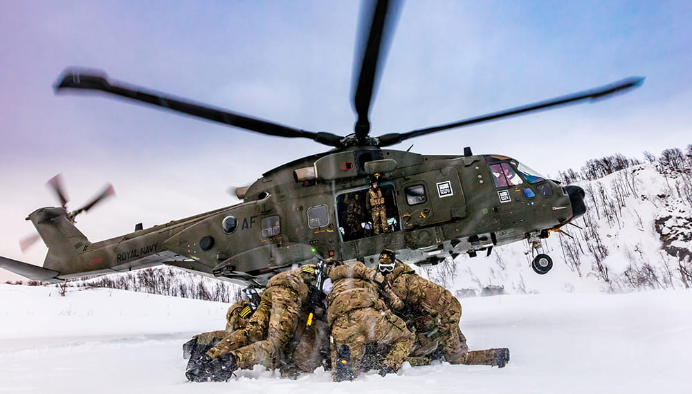 Merlin Mk3A helicopter lands on snowy ground with fatigue wearing Royal Marines ducked down beneath a purple grey sky