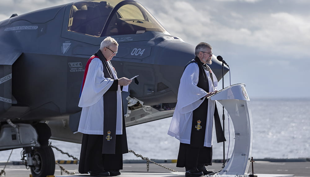 two robed royal navy chaplains give a service on deck with f35 jet in background