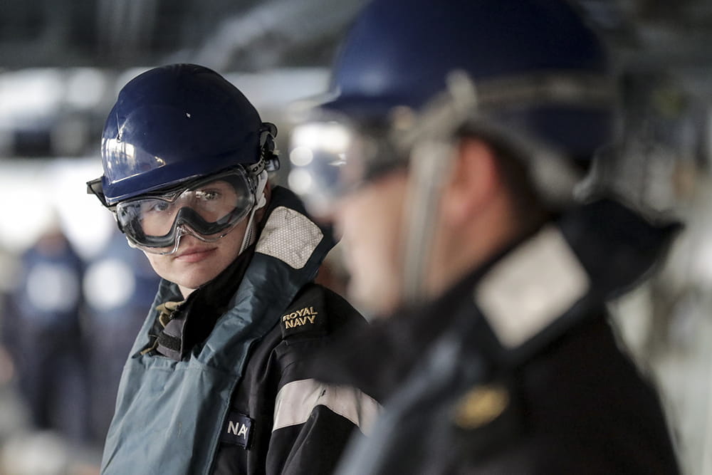 A Royal Navy sailor wearing a helmet and eye protection looks directly at the camera
