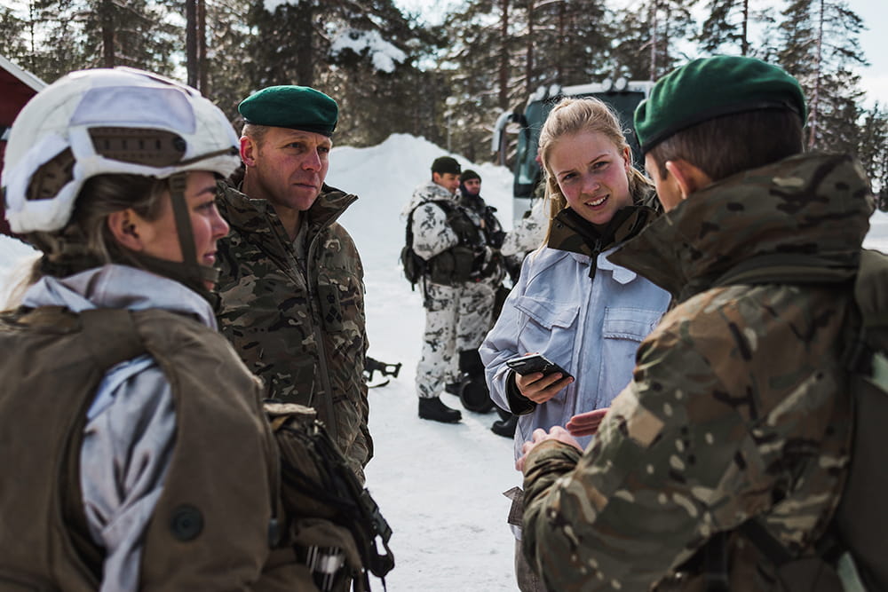 A group of people wearing cameo, green berets and helmets talk among themselves in the snowy outdoors