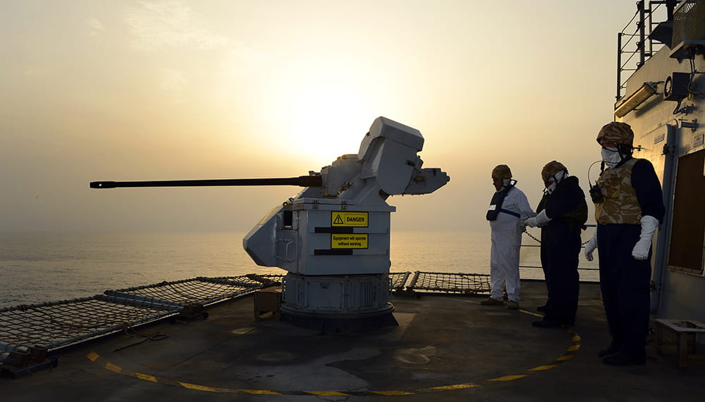 Three Royal Navy sailors stand beside a large gun onboard RFA Cardigan Bay while the sun sets
