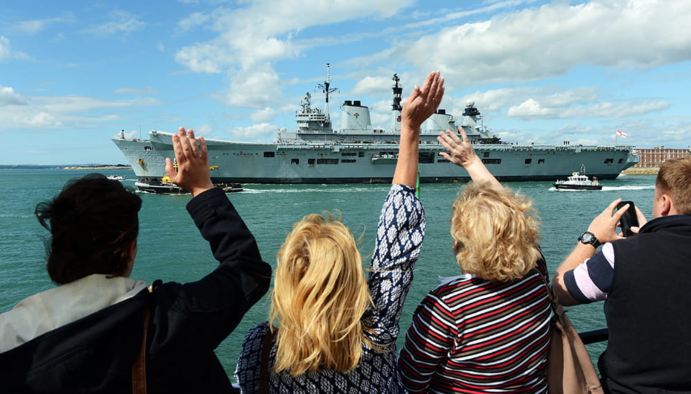 Friends and family wave towards a Royal Navy ship from the coast