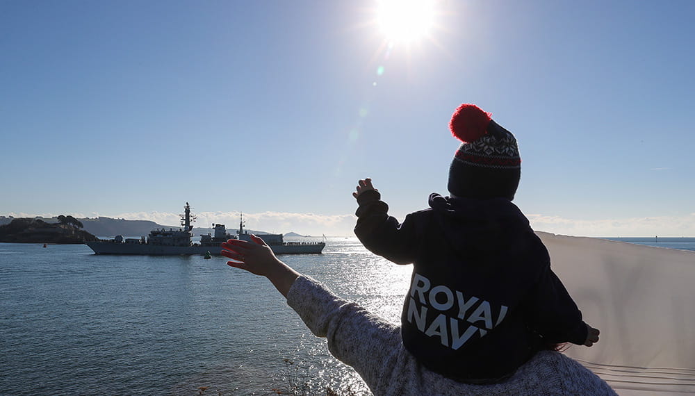 Small child with bobblehat on parent's shoulders waving off HMS Montrose