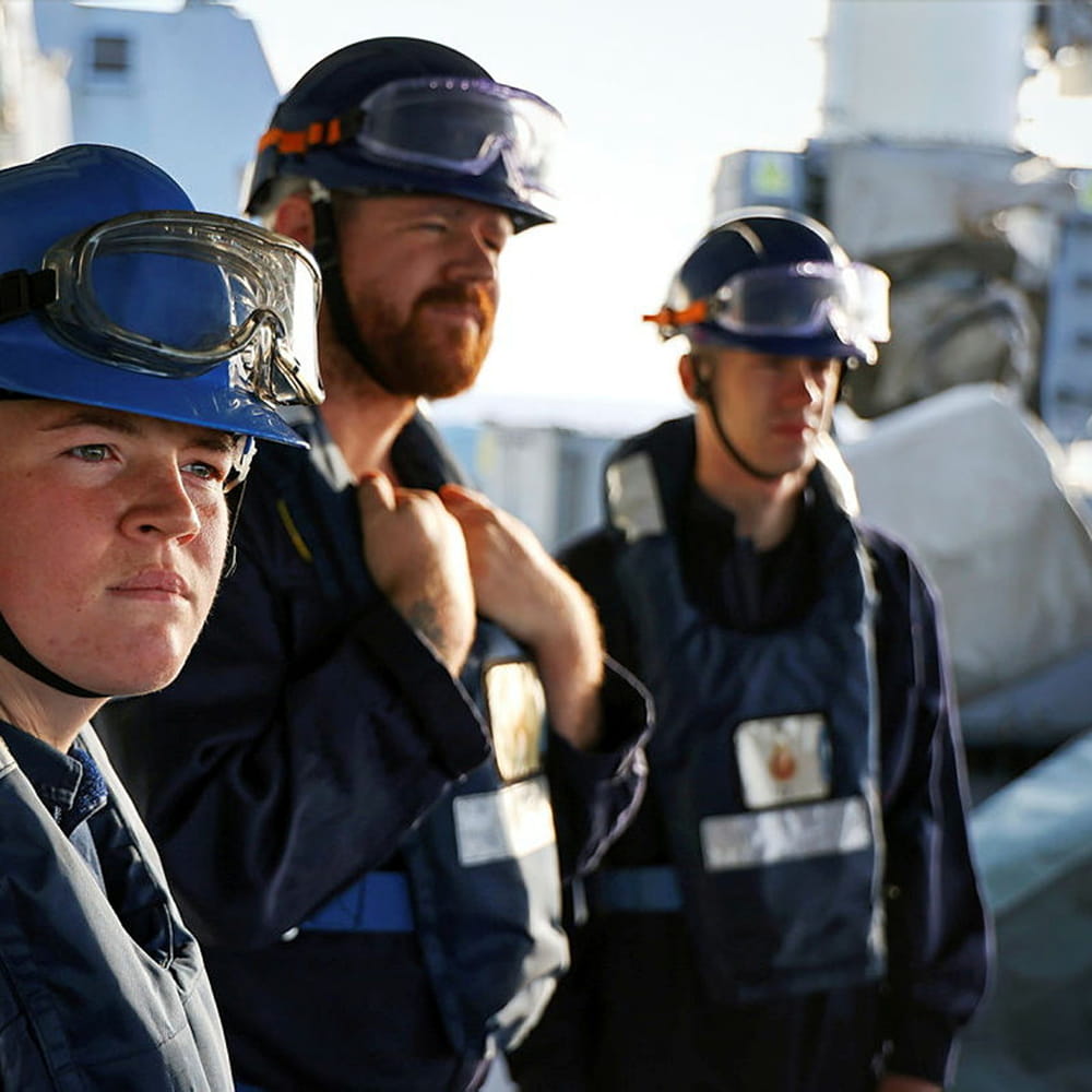 Three Royal Fleet Auxillary sailors in blue hard hats onboard the HMS Dragon