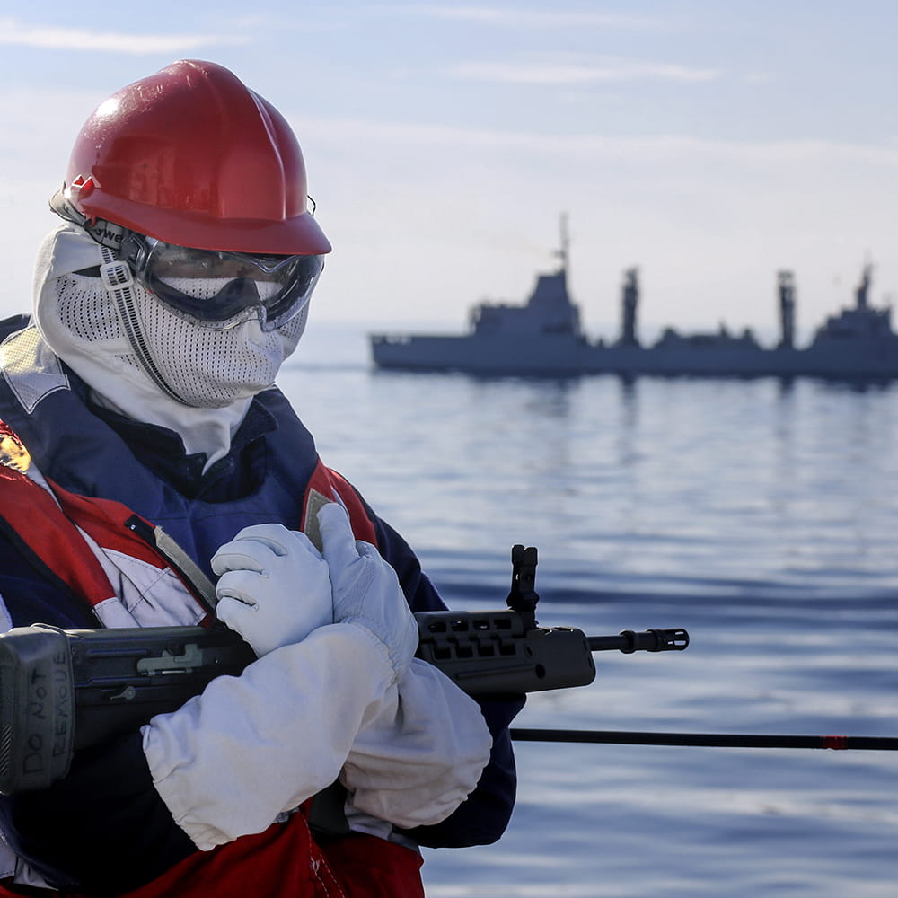 Armed Royal Navy sailor with a red hard hat, goggles and white face mask with a military ship in background