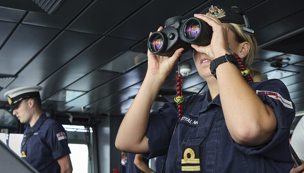 Royal Navy officer looks through binoculars from the bridge of a ship