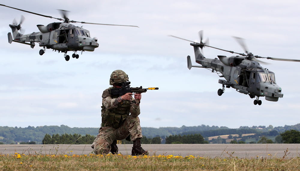 Royal Marine on one knee aims his gun while two two helicopters fly overhead